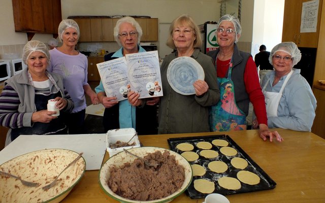 Onderskeidelik 3de en 4de van links is Suster Linda Meyer en Francia van Dyk met die sertifikaat en bord-embleem wat deur BADISA aan Gansbaai se Goue Hande toegeken is, saam met enkele lede by hulle wat besig is om pasteie te bak